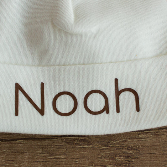 Close-up van een ecru babymutsje met de naam Noah in chocoladebruine letters op de omslag.