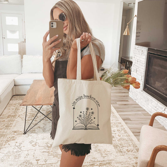 Vrouw poseert met canvas tote bag met de tekst “in mijn boeken-era” en bloemenillustratie, gevuld met tulpen in een moderne woonkamer.