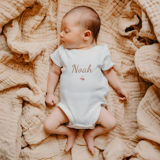 Baby liggend op een zachte beige deken draagt een wit rompertje met de naam Noah en een klein blush ballonnen ontwerp op de borst.