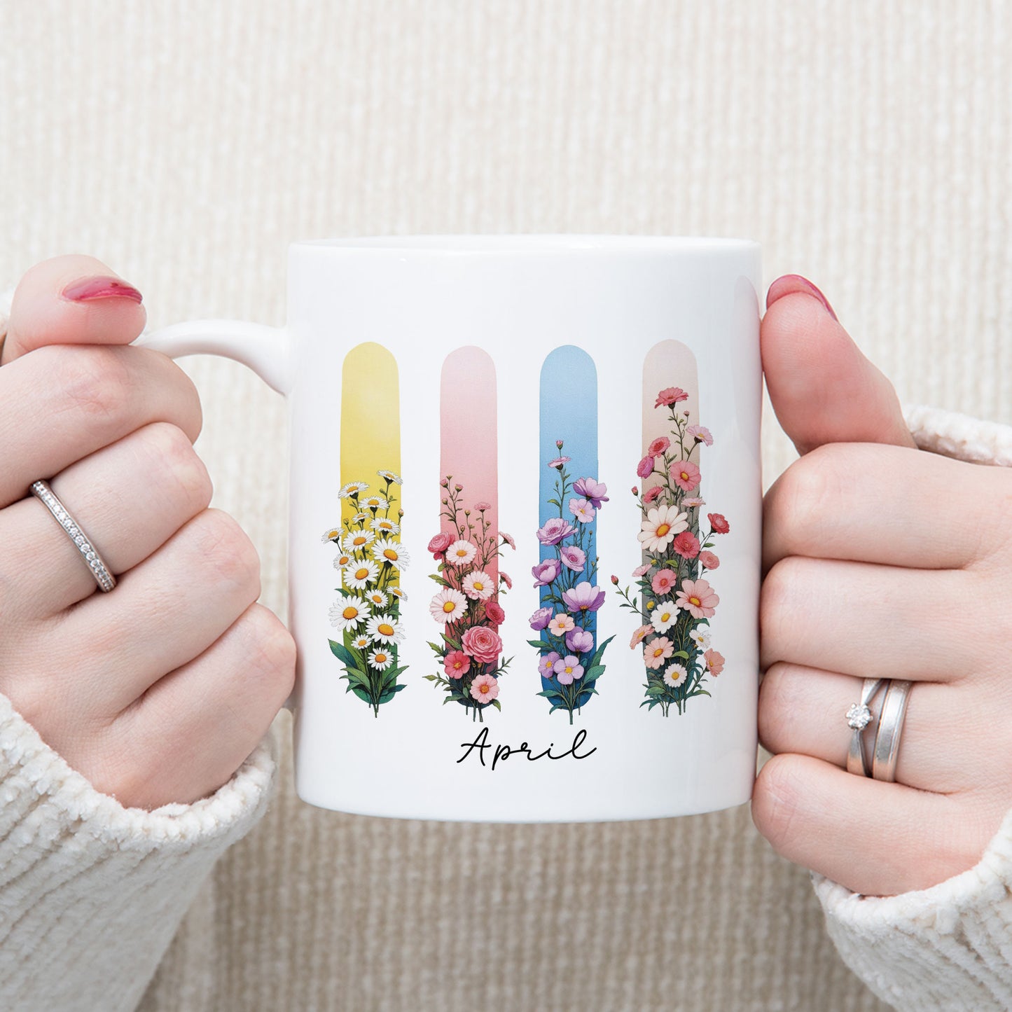 Persoon in een witte gebreide trui houdt een witte mok met de tekst April en bloemen in brush effect met beide handen vast.