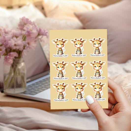 Persoonlijk stickervel met 9 giraf verjaardag stickers met de tekst 'Hoera Bella 6 jaar', vastgehouden in een hand voor een laptop en roze bloemen.