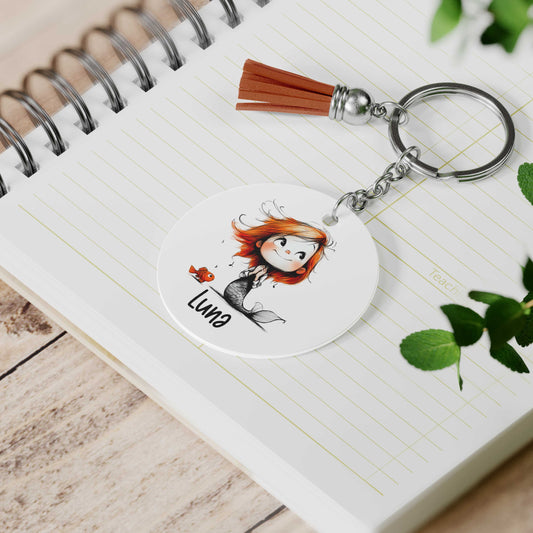 Gepersonaliseerde zeemeermin sleutelhanger met visje en voorbeeldnaam Luna liggend op een open notitieboek naast groene bladeren.