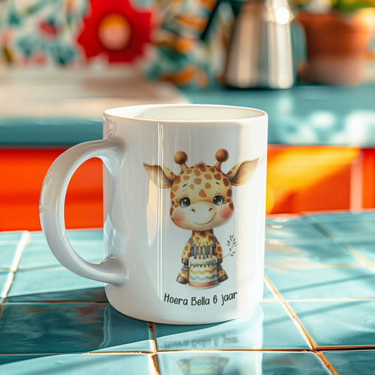 Witte keramische mok met giraf en verjaardagstaart, tekst ‘Hoera Bella 6 jaar’, staand op blauwe tegels in kleurrijke keuken.