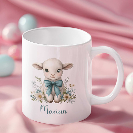 Witte mok met een schattige illustratie van een lammetje met blauwe strik en bloemen rondom, gepersonaliseerd met de naam Marian, gefotografeerd op een roze satijnen ondergrond met pastel paaseieren.