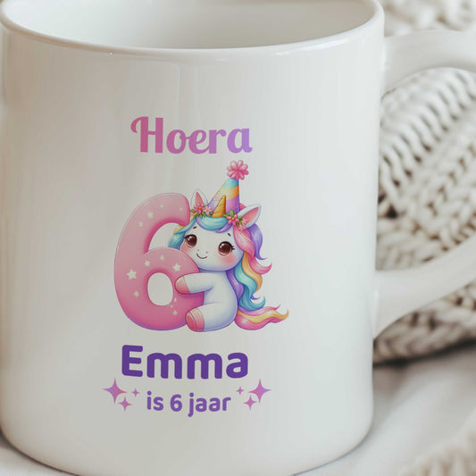 Close-up van een witte mok met een unicorn die een groot roze cijfer 6 vasthoudt en de tekst 'Hoera Emma is 6 jaar'.