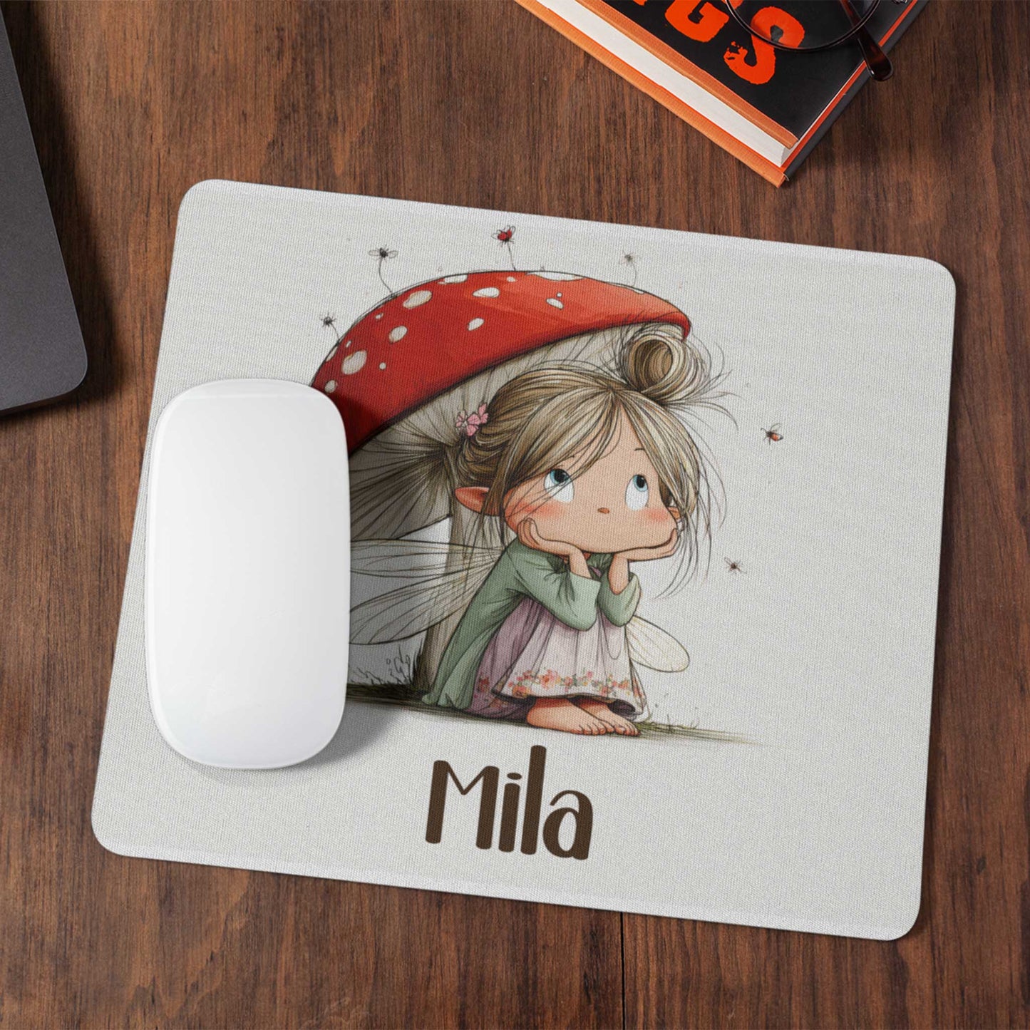 Muismat op een donker houten bureau met een illustratie van een fee onder een rode paddenstoel en de naam Mia.