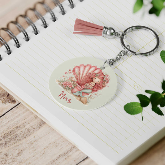 Ronde sleutelhanger met roze kwastje liggend op een notitieboek. De sleutelhanger toont een roze zeemeermin in een grote schelp, omringd door bloemen en schelpen, met de naam Noor onder de illustratie.