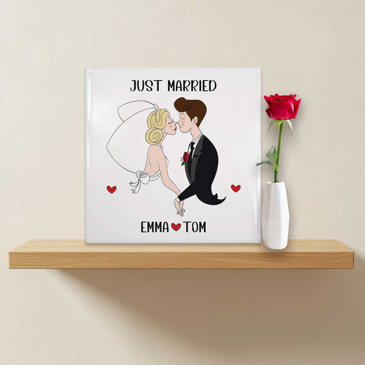 Cartoon trouwtegeltje op een houten plank met een witte vaas en rode roos ernaast, met de tekst 'Just Married' en namen Emma & Tom – illustratie van een bruidspaar dat elkaar kust.