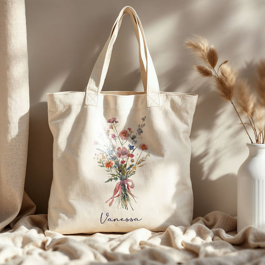 Totebag van katoen met boeket wilde bloemen en roze lint, staand op lichte stof naast een witte vaas