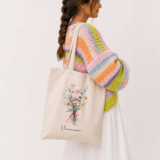 Vrouw met lange vlecht draagt een beige totebag met een boeket wilde bloemen in zachte pastelkleuren en de naam Vanessa. Ze draagt een kleurrijke gebreide trui in roze, oranje, groen en paars tinten.