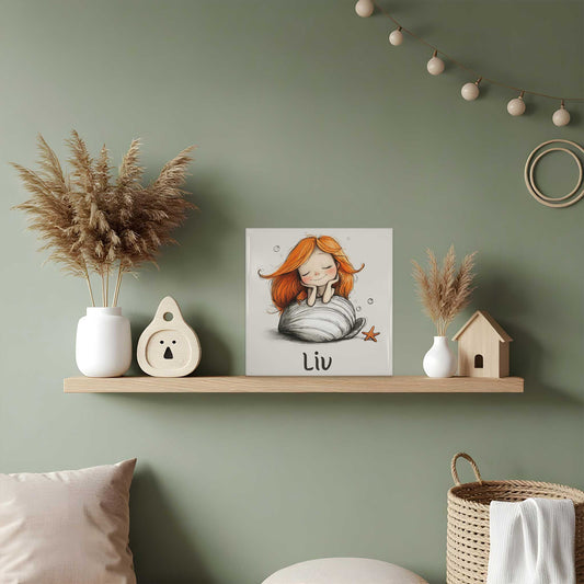 Keramisch tegeltje met een zeemeermin met lang oranje haar die dromend op een grote witte schelp rust. Onder de illustratie staat de naam Liv. Het tegeltje staat op een houten plank tegen een groene muur.