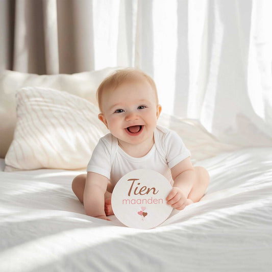 Lachende baby in wit rompertje op bed met mijlpaalschijfje met tekst tien maanden in de handen