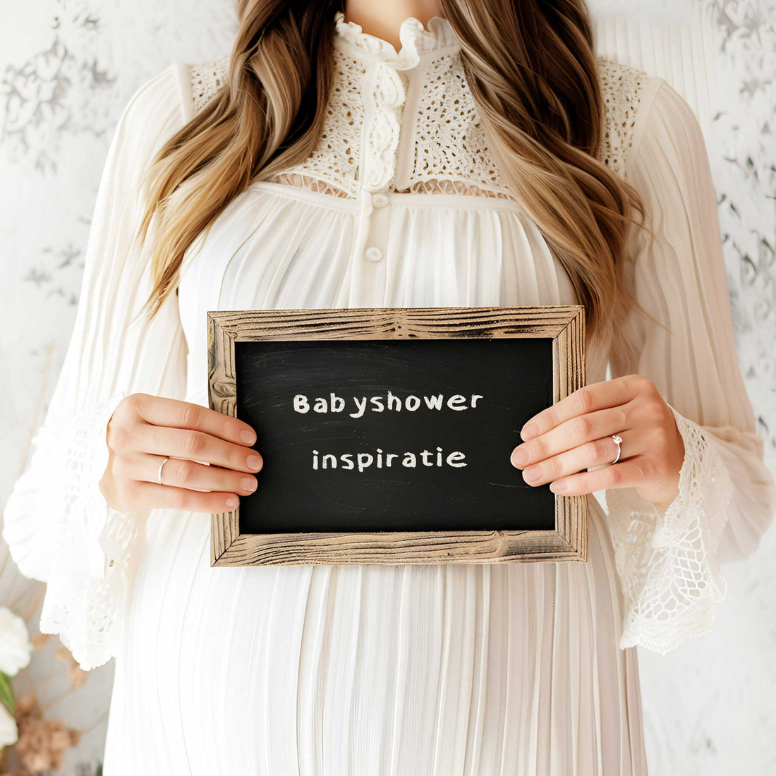 Zwangere vrouw in een witte jurk houdt een klein krijtbord vast met de tekst “Babyshower inspiratie” in een zachte neutrale setting.