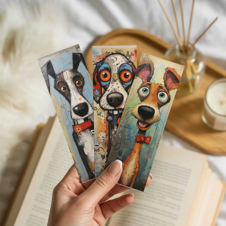 Hand houdt drie boekenleggers met kleurrijke, artistieke hondenillustraties vast boven een open boek in een lichte, warme setting.