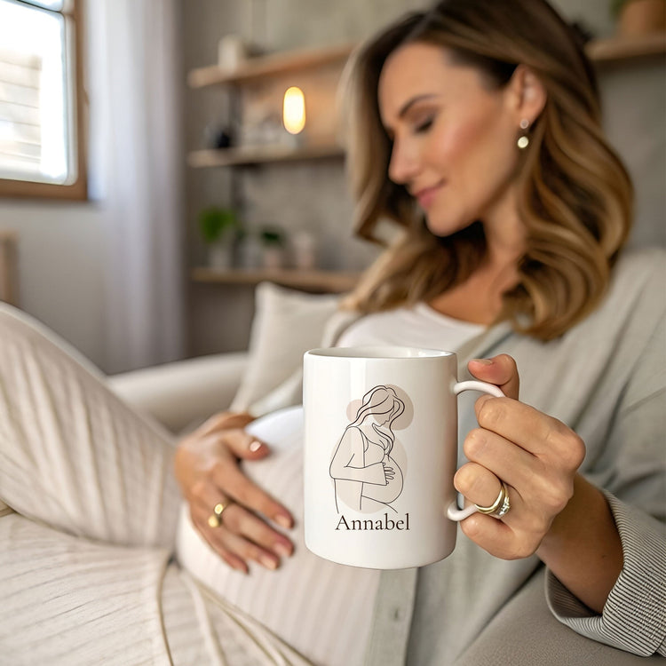 Zwangere vrouw ontspant op de bank met een gepersonaliseerde witte mok met minimalistische lijntekening van een zwangere vrouw en de naam Annabel. Sfeervolle lifestylefoto in zachte neutrale tinten, passend bij zwangerschap en babyshower cadeau inspiratie