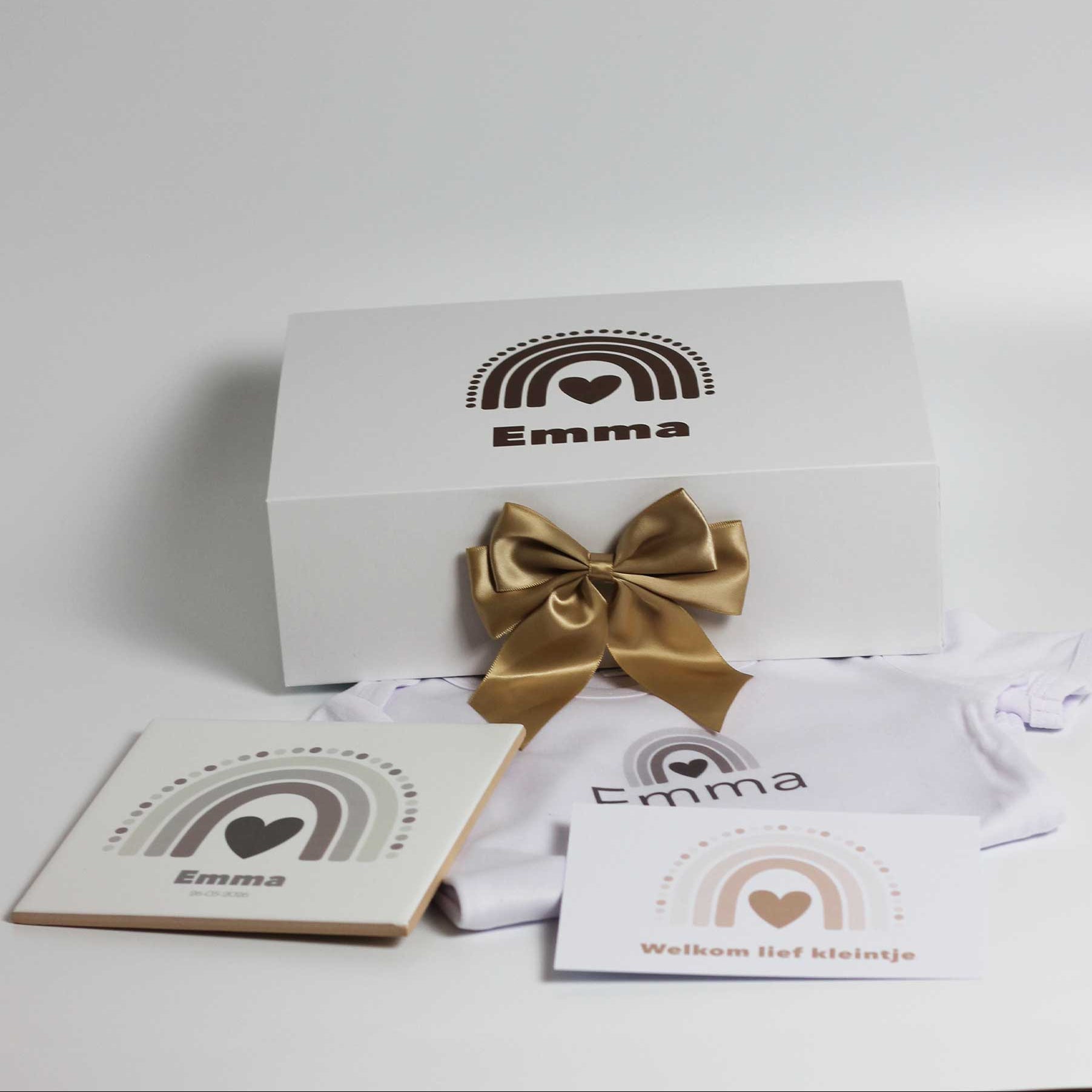 Witte cadeaudoos met naam Emma en regenboogprint met gouden strik, met daarvoor een wit babyrompertje met naam, keramisch tegeltje en kaartje op een witte achtergrond