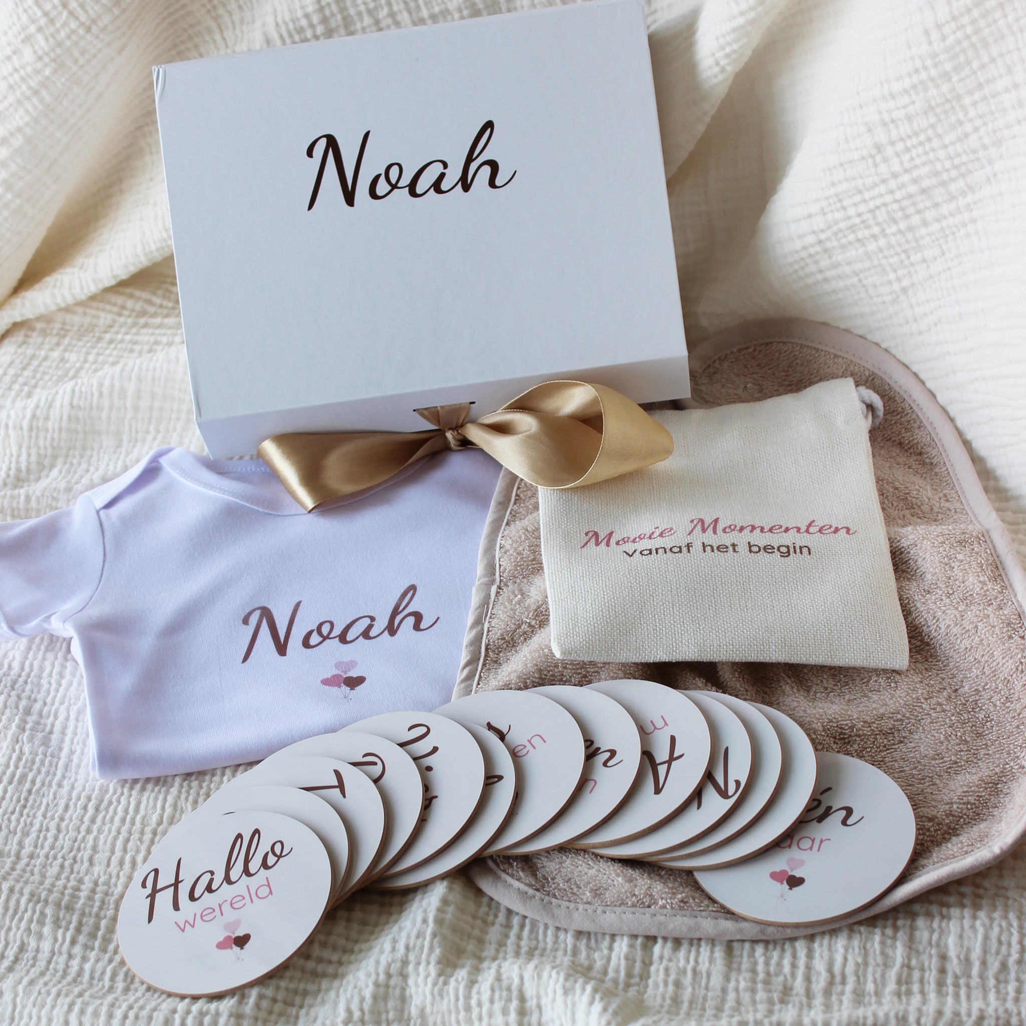 Baby giftbox met wit rompertje met naam Noah, beige slabbetje, mijlpaalschijfjes en linnen zakje op een beige mousseline doek