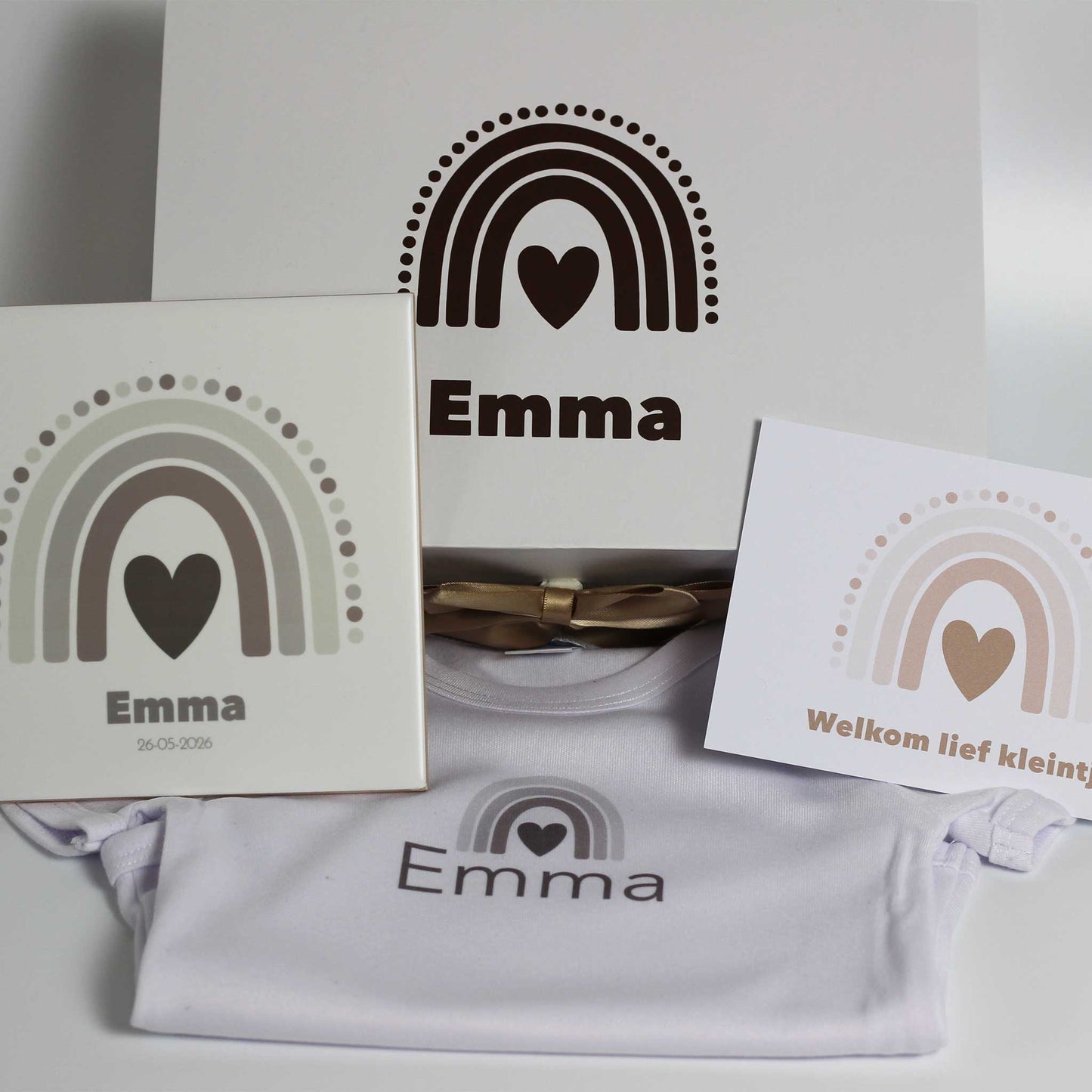 Witte baby giftbox met naam Emma en regenboogprint op de doos, met daarvoor een wit babyrompertje met naam, een keramisch tegeltje met naam en geboortedatum en een kaartje met regenboog op een witte achtergrond