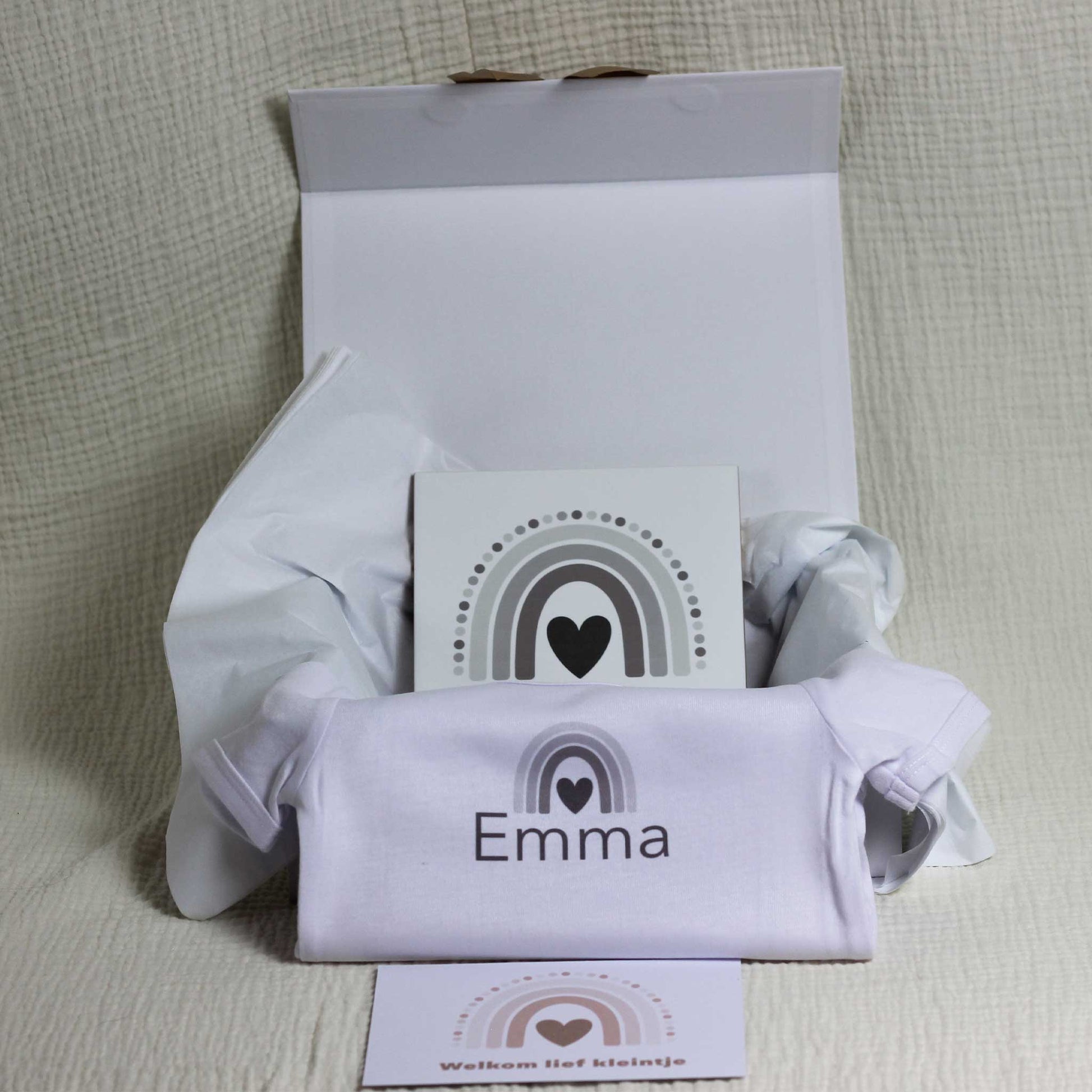 Geopende witte cadeaudoos op een zachte beige stof met wit zijdepapier, waarin een keramisch tegeltje met regenboog zichtbaar is, met daarvoor een wit babyrompertje met naam Emma en een kaartje met regenboog en de tekst “Welkom lief kleintje”