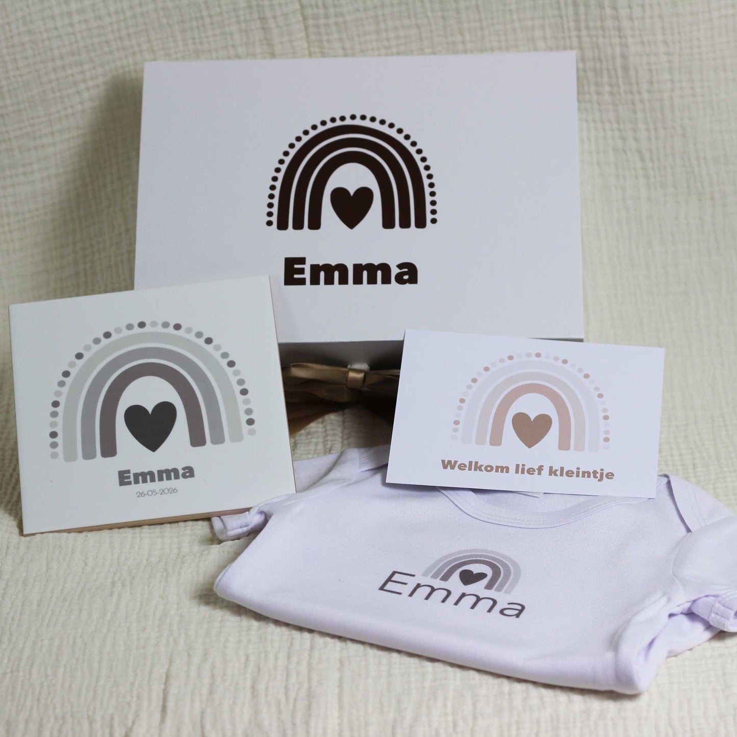 Witte cadeaudoos met regenboog en naam Emma op een zachte beige stof, met daarvoor een keramisch tegeltje met naam en geboortedatum, een kaartje met regenboog en de tekst “Welkom lief kleintje” en een wit babyrompertje met naam