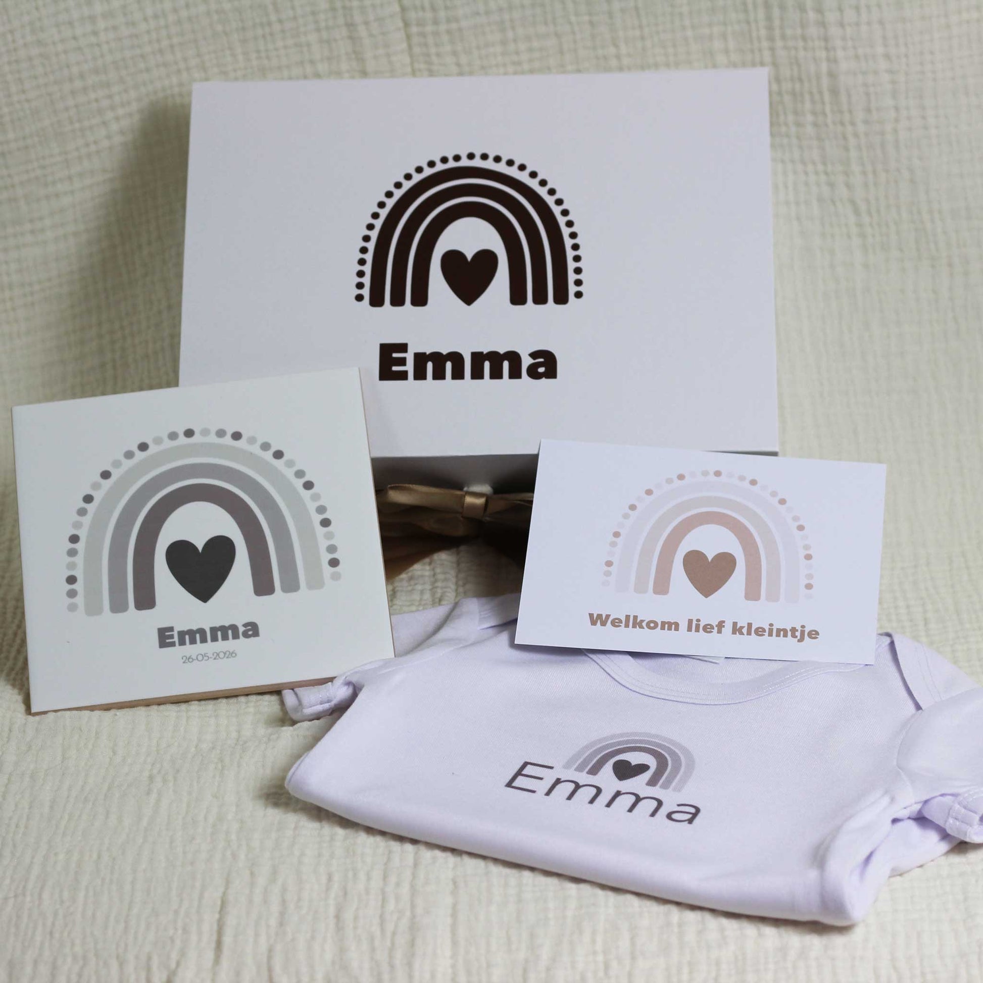 Witte cadeaudoos met regenboog en naam Emma op een zachte beige stof, met daarvoor een keramisch tegeltje met naam en geboortedatum, een kaartje met regenboog en de tekst “Welkom lief kleintje” en een wit babyrompertje met naam