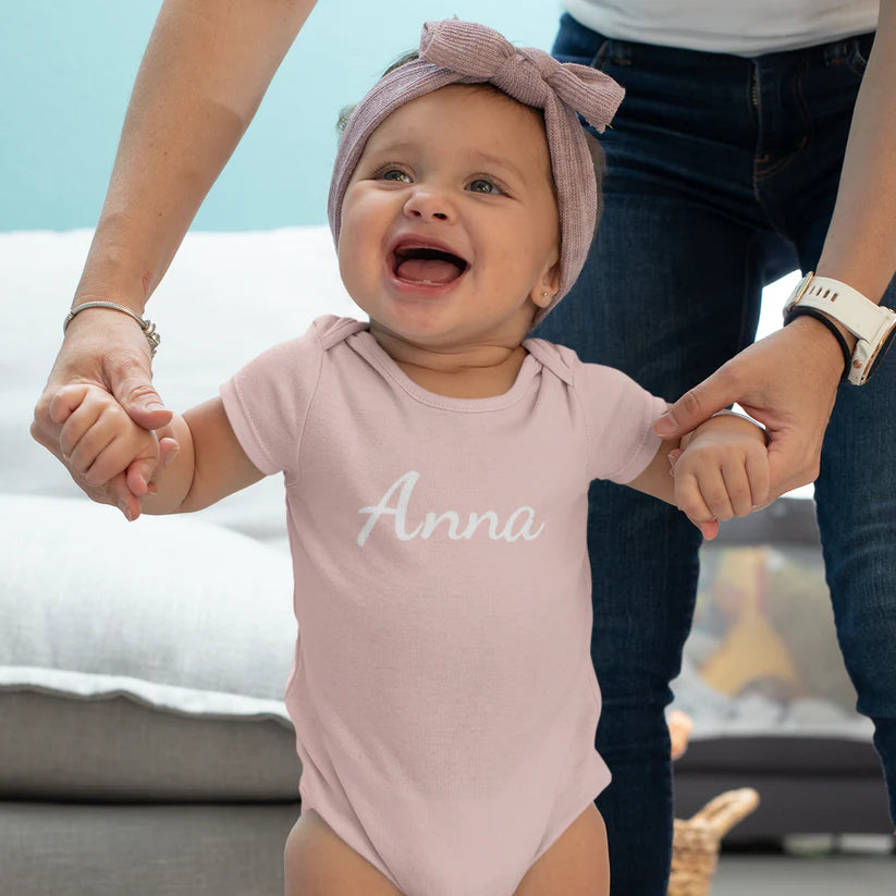 Baby draagt roze rompertje met naam Anna terwijl een volwassene haar handen vasthoudt