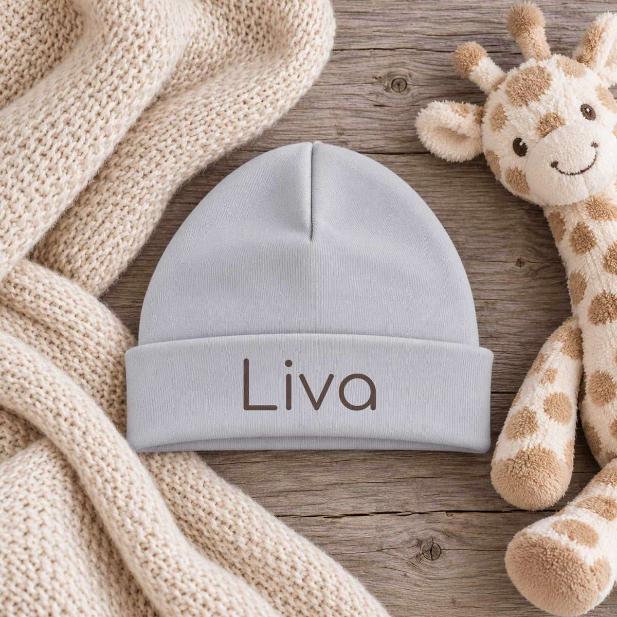 Sandstone Grey babymutsje op een grijze houten tafel met een ecru gebreide doek en zacht girafje ernaast