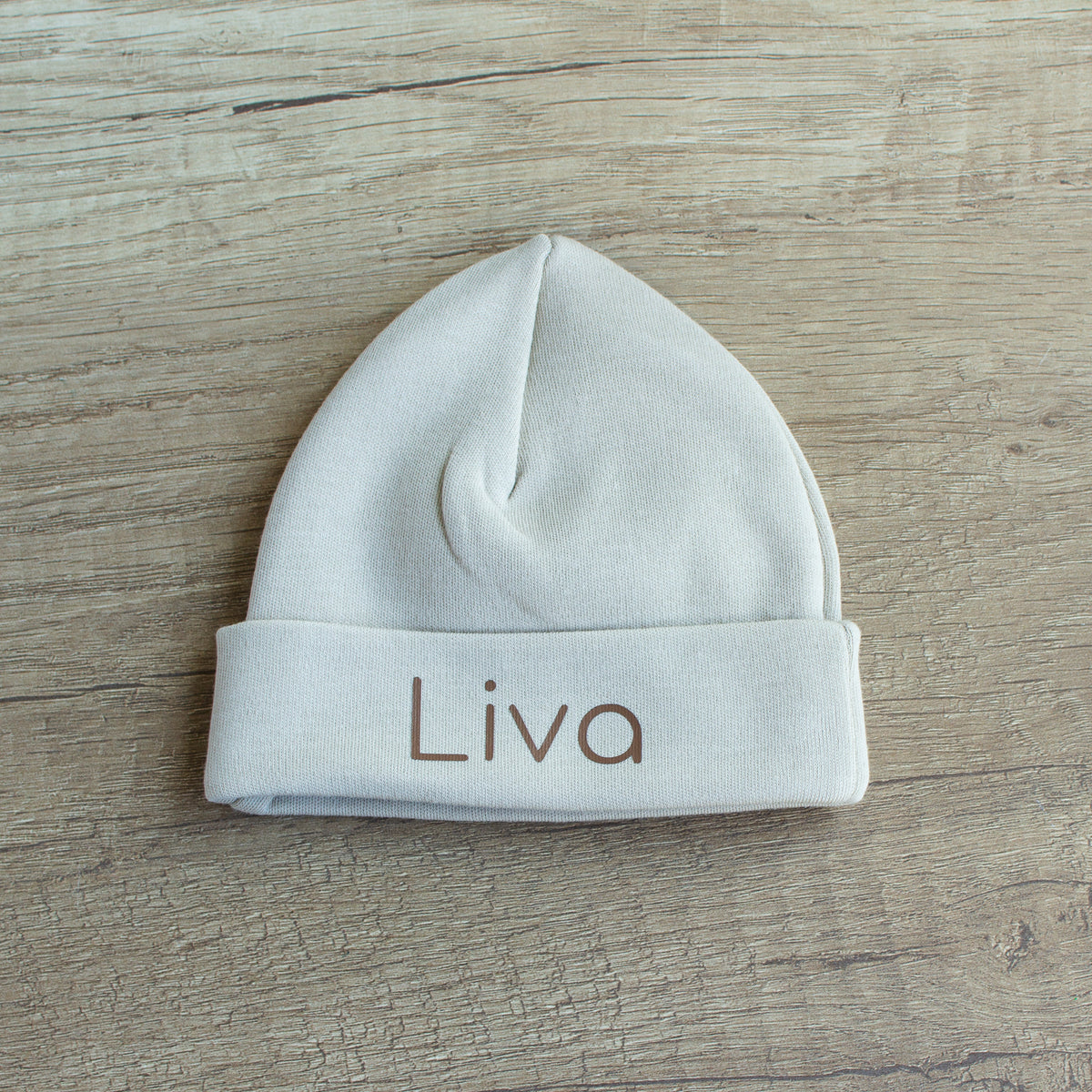 Gepersonaliseerd newborn babymutsje in Sandstone Grey met de naam Liva in chocoladebruin, liggend op een houten tafel.