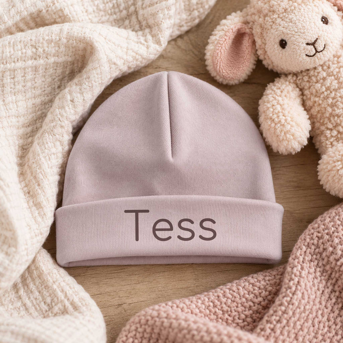 Pinkstone babymutsje met de naam Tess op een houten tafel met zachte ecru stoffen en een lammetje ernaast