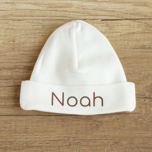 Ecru babymutsje maat 50/56 met de naam Noah in chocoladebruine letters, liggend op een houten tafel.