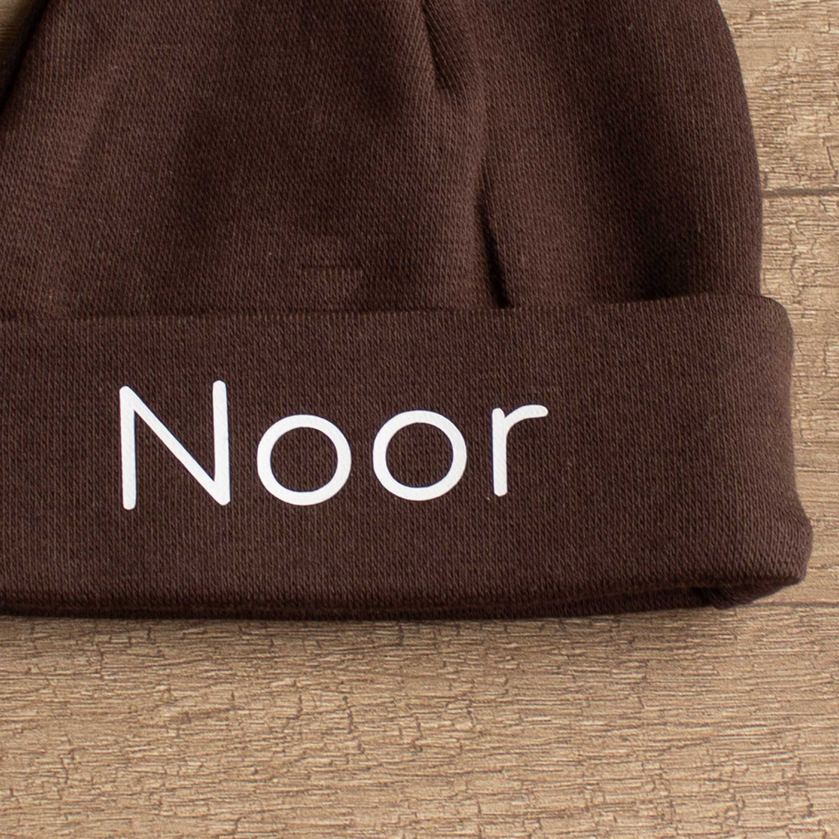 Close-up van chocoladebruin babymutsje met de naam Noor in wit, met zicht op de katoenen stof en omgeslagen rand.