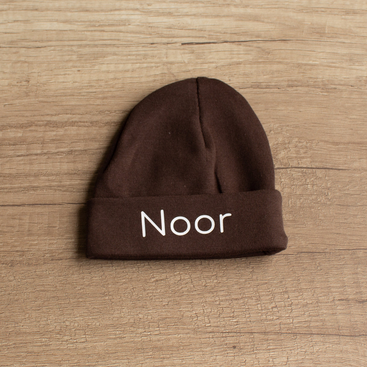 Chocoladebruin babymutsje van 100% katoen met witte naam Noor, liggend op een lichte houten ondergrond.