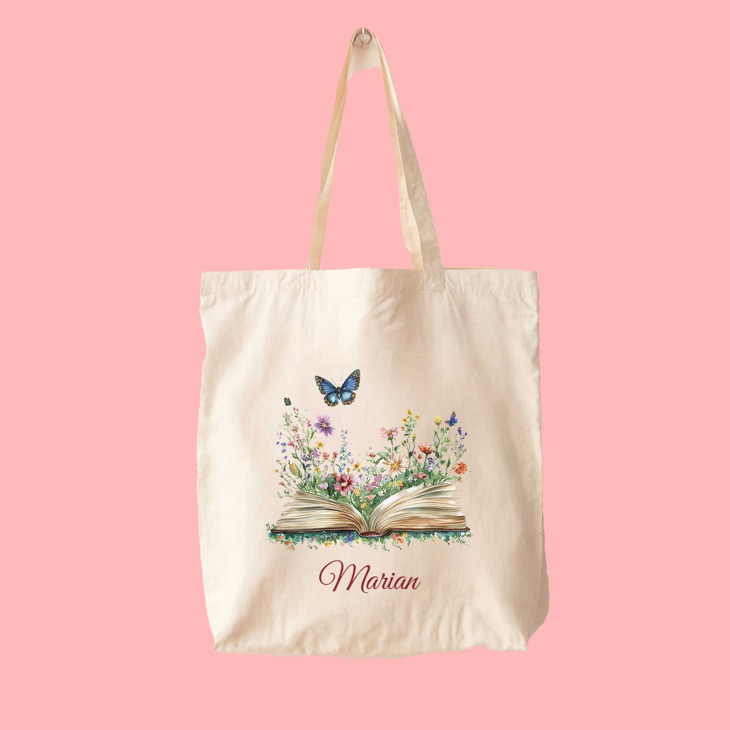 Canvas tote bag met kleurrijke bloemen die uit een open boek groeien, een vlinder erboven en de naam Marian gedrukt eronder, op een roze achtergrond.