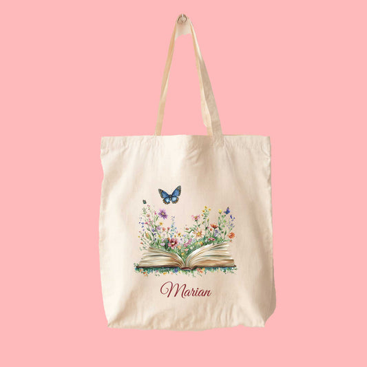 Canvas tote bag met kleurrijke bloemen die uit een open boek groeien, een vlinder erboven en de naam Marian gedrukt eronder, op een roze achtergrond.