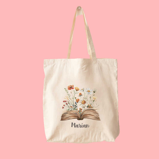Canvas tote bag met illustratie van open boek en veldbloemen in aardetinten, gepersonaliseerd met de naam Marian, op een roze achtergrond.
