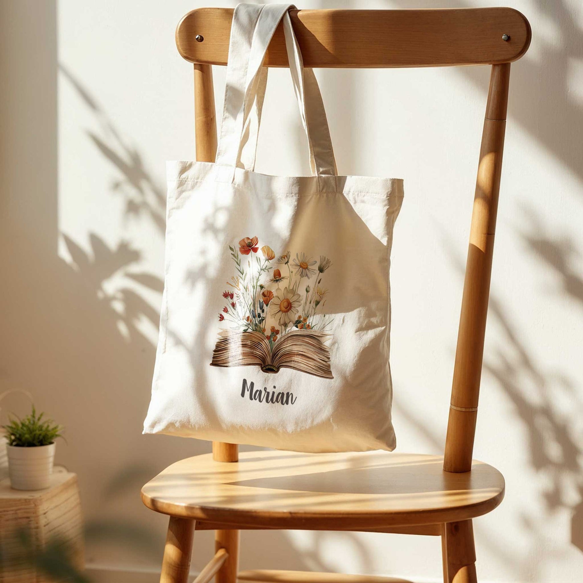 Gepersonaliseerde canvas tote bag met veldbloemen en naam "Marian", hangend aan een houten stoel in natuurlijk zonlicht.