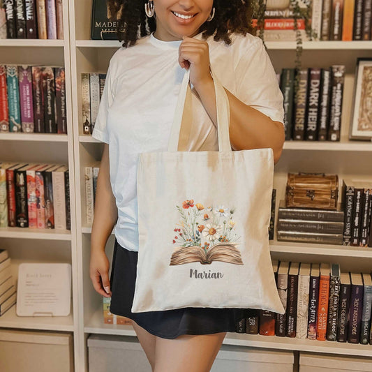 Vrouw poseert met gepersonaliseerde tote bag bedrukt met open boek en veldbloemen, in een boekenkamer vol romans.