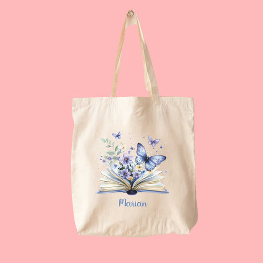 Canvas tote bag met een magische illustratie van blauwe bloemen en vlinders die uit een open boek komen, gepersonaliseerd met naam Marian.