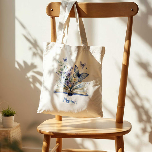 Gepersonaliseerde tote bag met blauwe vlinders en bloemen, hangend aan een houten stoel in warm natuurlijk licht.