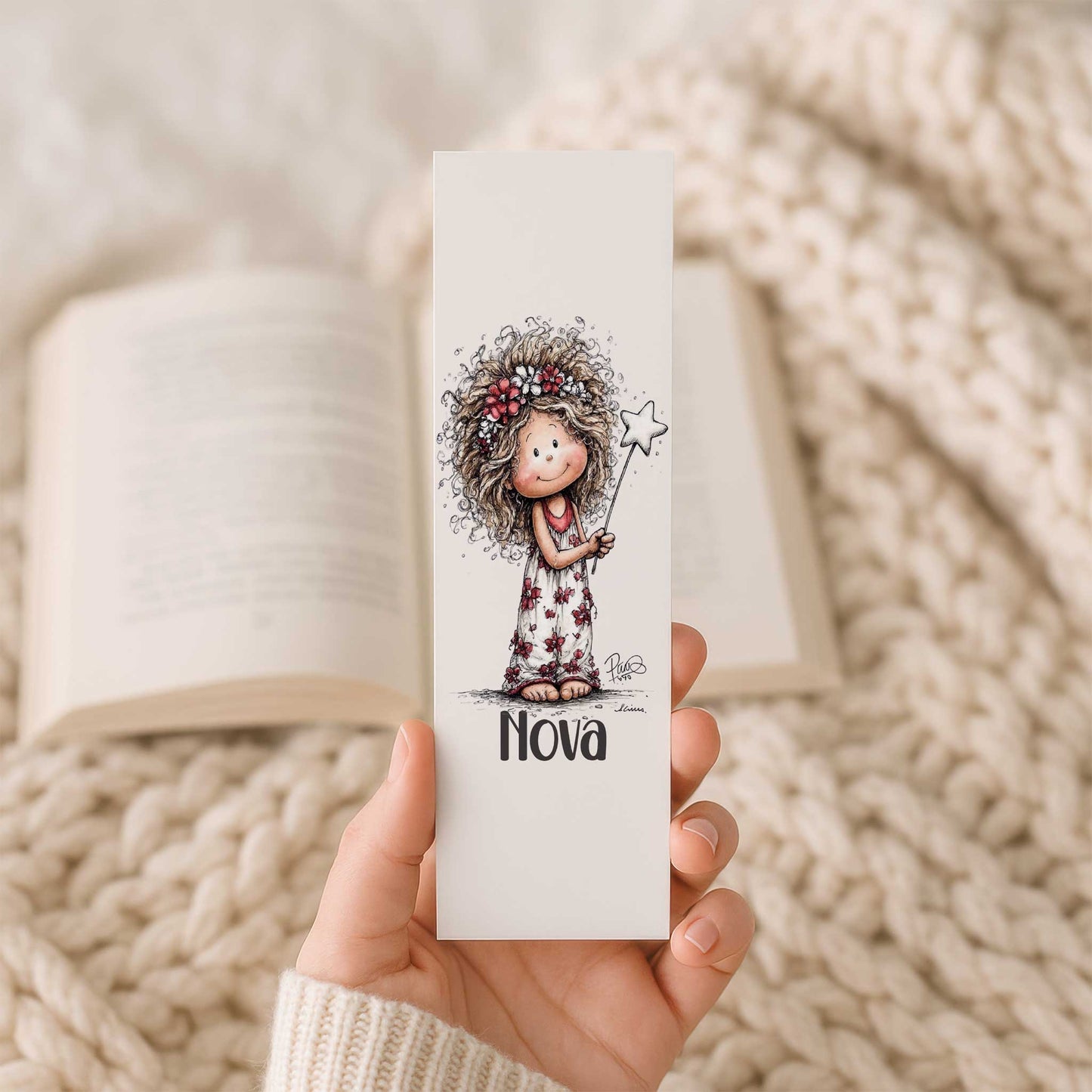 Hand houdt een boekenlegger met naam Nova en illustratie van een fee met bloemenkrans en toverstaf voor een open boek