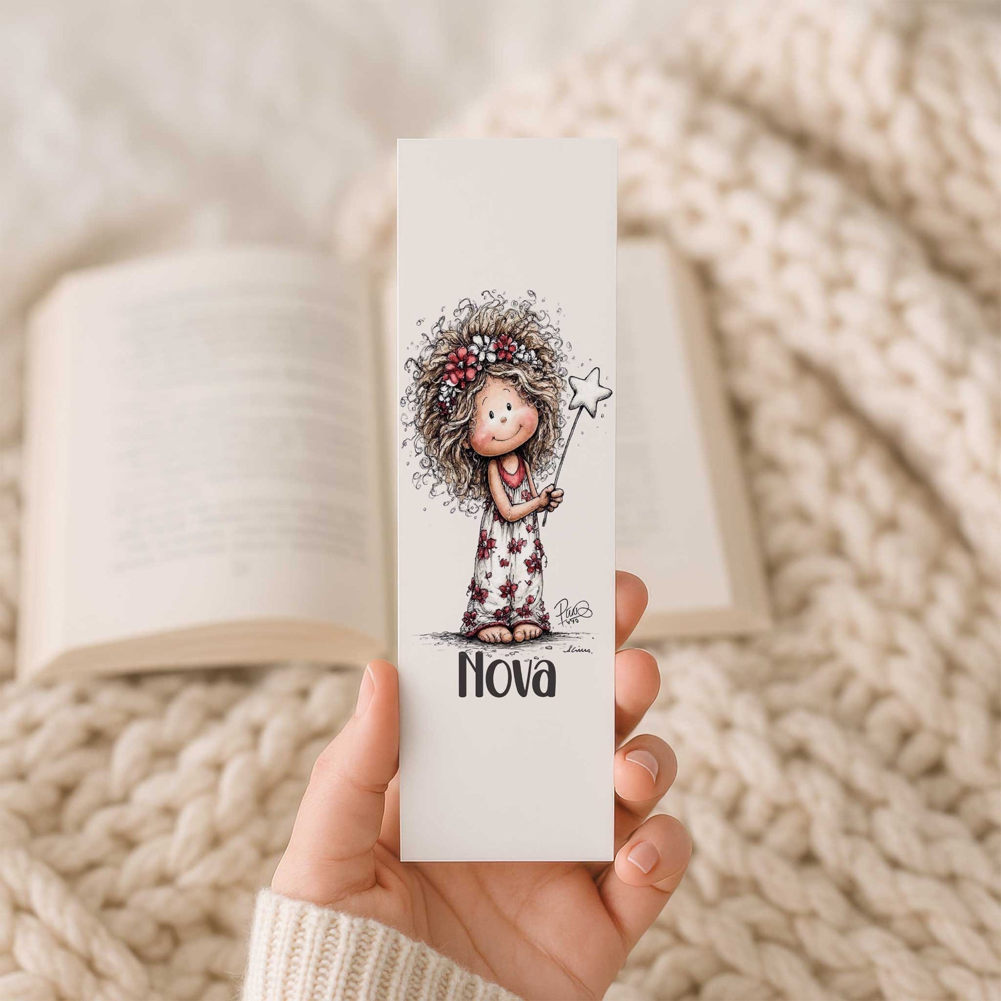 Hand houdt een boekenlegger met naam Nova en illustratie van een fee met bloemenkrans en toverstaf voor een open boek