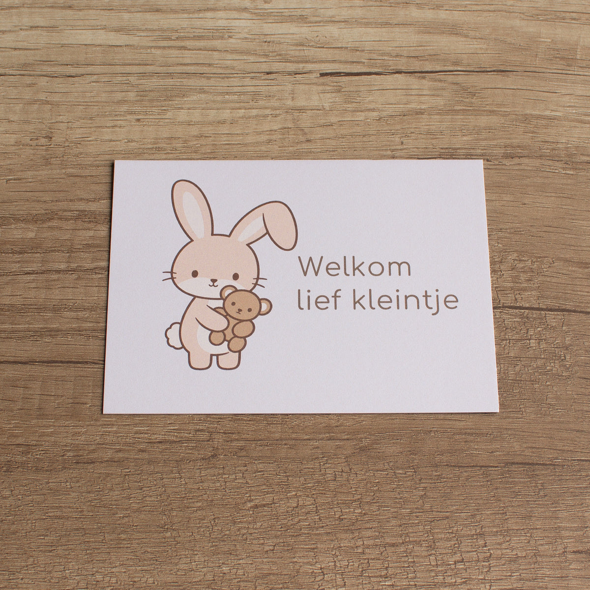 Bijpassend geboortekaartje met illustratie van een konijntje met beertje en de tekst Welkom lief kleintje, liggend op een houten tafel.