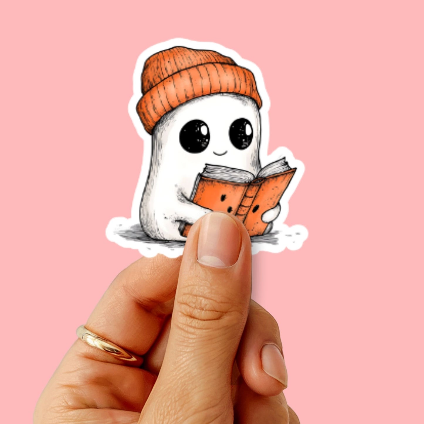 Sticker met cozy ghost wordt vastgehouden voor een roze achtergrond; spookje leest een oranje boek en draagt een warme muts.
