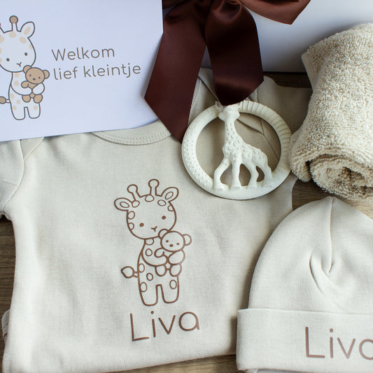 Close-up van de naturel babycadeauset met een ecru romper en muts, beide gepersonaliseerd met de naam Liva. Op de romper staat een getekend girafje met een beertje. Bovenop ligt een crèmekleurige bijtring in de vorm van een giraf. Rechts ligt een opgerold ecru spuugdoekje en bovenaan is een chocoladebruine strik zichtbaar. Links staat een kaart met een girafje en de tekst ‘Welkom lief kleintje’.