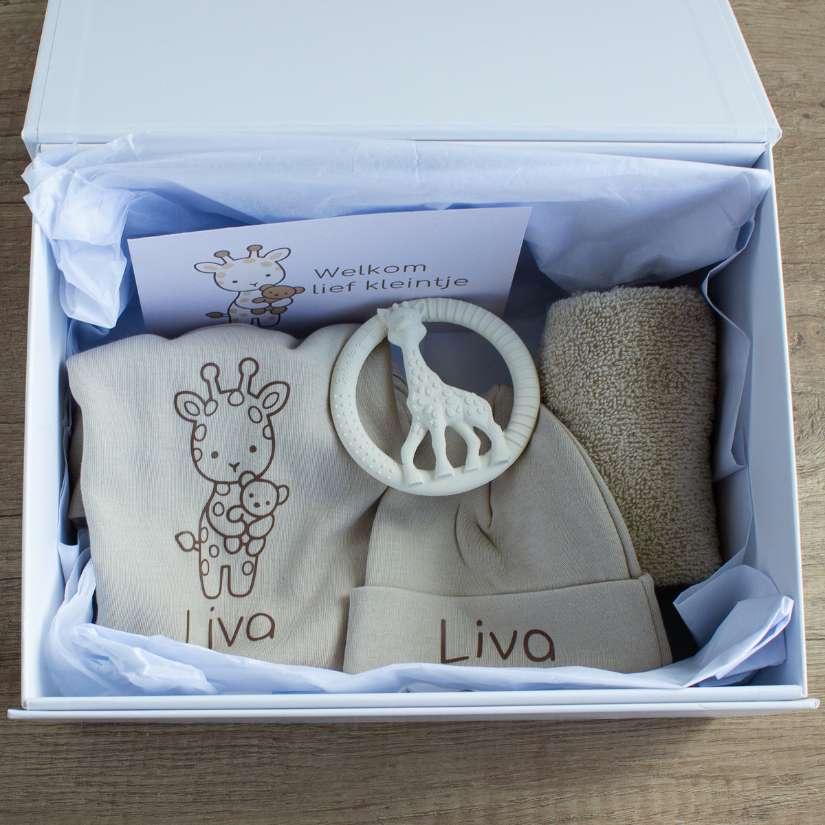 Geopende witte luxe cadeaudoos met een ecru romper en muts, beide gepersonaliseerd met de naam Liva. Op de romper staat een getekend girafje met een beertje. Bovenop ligt een crèmekleurige bijtring in de vorm van een giraf en rechts een opgerold ecru spuugdoekje. Achterin staat een kaart met een girafje en de tekst ‘Welkom lief kleintje’. De cadeauset ligt in wit vloeipapier.
