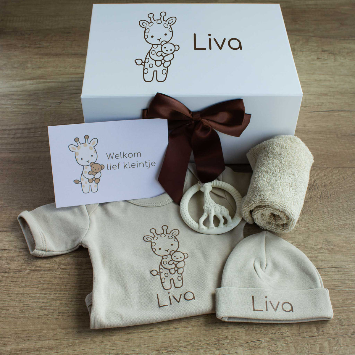 Naturel babycadeauset met een gepersonaliseerde ecru romper en muts met de naam Liva. Achter de kleding staat een witte cadeaudoos met een getekend girafje en de naam Liva, versierd met een chocoladebruine strik. Bij de set liggen een crèmekleurige bijtring in de vorm van een giraf, een opgerold ecru spuugdoekje en een kaart met een girafje en de tekst ‘Welkom lief kleintje’.