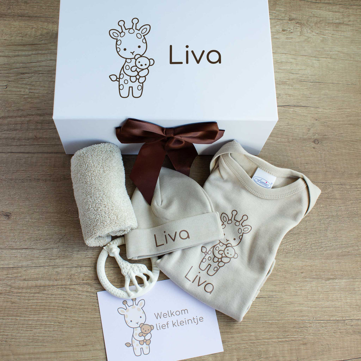 Luxe babycadeauset in zandkleur met een witte magneetdoos, gepersonaliseerd met een girafje en de naam Liva. Voor de doos liggen een romper en muts met naam, een opgerold spuugdoekje, een crèmekleurige bijtring in de vorm van een giraf en een kaart met de tekst ‘Welkom lief kleintje’. De witte cadeaudoos is afgewerkt met een chocoladebruine satijnen strik.