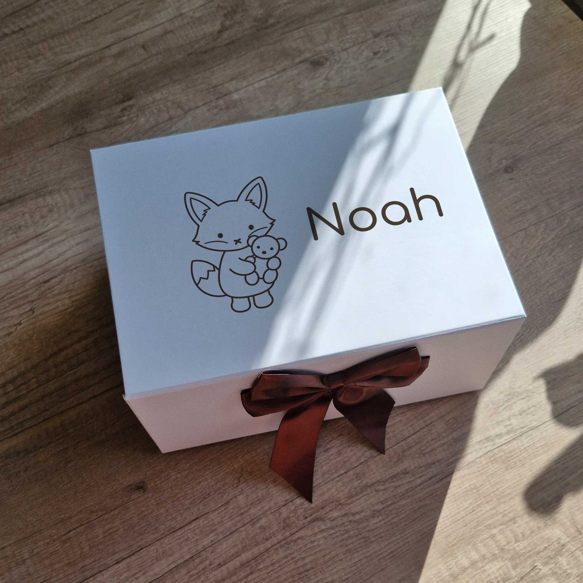 Witte luxe baby giftbox gepersonaliseerd met een vosje en de naam Noah, afgewerkt met een chocoladebruine satijnen strik. De doos staat op een houten ondergrond in natuurlijk zonlicht.
