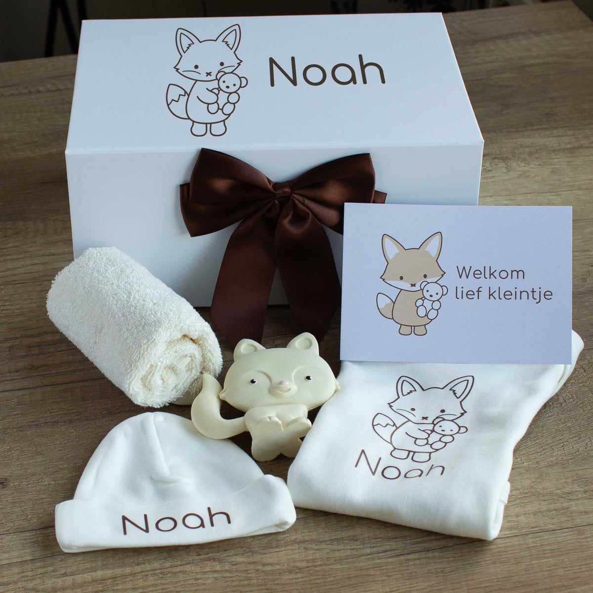 Luxe babycadeauset met vosje in ecru, met een gepersonaliseerde romper en muts met de naam Noah, een zacht ecru spuugdoekje, een crèmekleurig vosje als bijtspeeltje en een kaart met de tekst ‘Welkom lief kleintje’. Achter de cadeauset staat de witte gepersonaliseerde cadeaudoos met vosillustratie en de naam Noah, afgewerkt met een chocoladebruine satijnen strik.