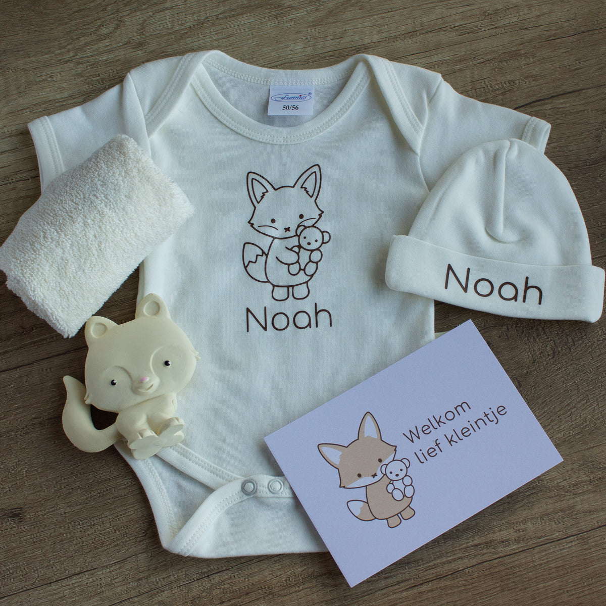 Inhoud van de luxe baby giftbox met vosje in ecru: gepersonaliseerd rompertje en mutsje met de naam Noah en vosillustratie, zacht ecru spuugdoekje, crèmekleurig vosje als bijtspeeltje en een kaart met de tekst ‘Welkom lief kleintje’, gefotografeerd op een houten ondergrond.